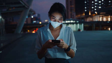 Young Asia businesswoman in fashion clothes wearing face mask using smart phone typing text message while stand outdoor in urban city at night.の写真素材