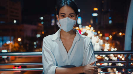 Successful young Asia businesswoman in fashion office clothes wear medical face mask smiling and looking at camera while happy stand alone outdoors in urban modern city night. Business on go concept.の写真素材