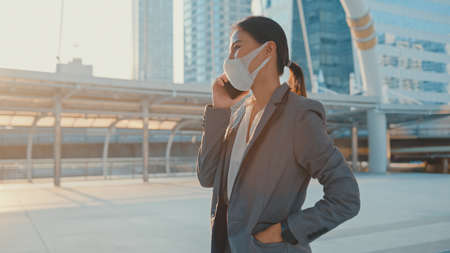 Young Asia businesswoman in fashion office clothe wear medical face mask talk via phone while walk alone outdoor in urban city.の写真素材