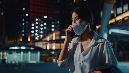 Serious dissatisfied upset young Asia businesswoman wear medical face mask talk via phone while walk alone outdoor in urban city night.の写真素材