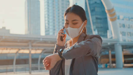 Young Asia businesswoman in fashion office clothe wear medical face mask talk via phone while walk alone outdoor in urban city.の写真素材