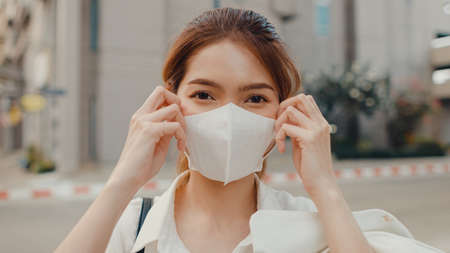 Successful young Asia businesswoman in fashion office clothes wear medical face mask smiling and looking at camera while happy standing alone outdoors in urban modern city. Business on the go concept.の写真素材