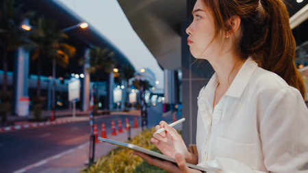 Successful young Asia businesswoman in fashion office clothes using smart pen technology for write on digital tablet while sit alone outdoors in urban modern city at night. Business on the go concept.の写真素材