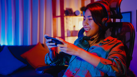 Happy asia girl gamer wear headphone competition play video game online with smartphone colorful...