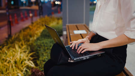 Successful young Asia businesswoman in fashion office clothes using laptop work remotely while sitting alone outdoors in urban modern city in the evening. Business on the go concept.の写真素材