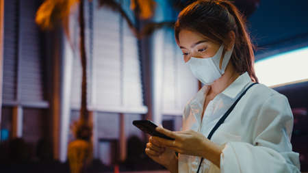 Young Asia businesswoman in fashion office clothes wearing medical face mask using smart phone typing text message while stand outdoors in urban modern city at night. Business on the go concept.の写真素材