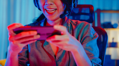 Happy asia girl gamer wear headphone competition play video game online with smartphone colorful...