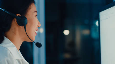 Millennial Asia young call center agent or customer support service executive using computer and microphone headset working technical support in late night office. Telemarketing or sales job concept.の写真素材