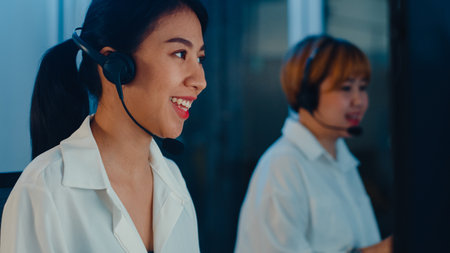 Millennial Asia young call center team or customer support service executive using computer and microphone headset working technical support in late night office. Telemarketing or sales job concept.の写真素材