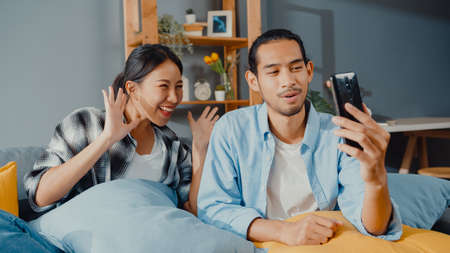 Happy young asian couple man and woman sit couch use smartphone video call with friends and family in living room at home. Stay at home quarantine, Social distancing, Young married concept.の写真素材