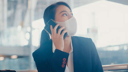 Smart asian business girl wear suit sitting with suitcase in bench call smart phone talk with partner wait for flight at airport. Business travel commuter in covid pandemic, Business travel concept.の写真素材
