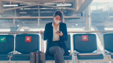 Asian business lady traveller wear suit sitting with suitcase and use smart phone chat message in bench wait for flight at airport. Business travel commuter in covid pandemic, Business travel concept.の写真素材