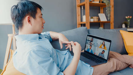 Young Asia businessman using laptop talk to colleagues about plan in video call meeting while work from home at living room. Self-isolation, social distancing, quarantine for   virus prevention.の写真素材