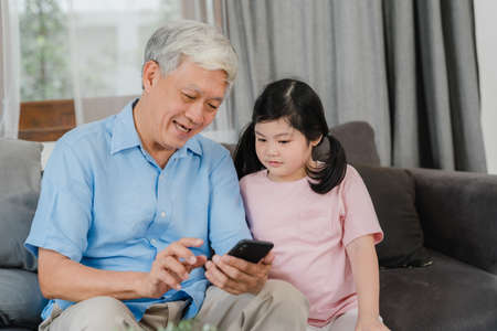 Asian grandparents and granddaughter using mobile phone at home. Senior Chinese, grandpa and kid happy spend family time relax with young girl checking social media, lying on sofa in living room.の写真素材