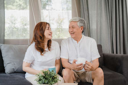 Asian senior couple relax at home. Asian Senior Chinese grandparents, husband and wife smile talking and drinking coffee while lying on sofa in living room at home concept.の写真素材