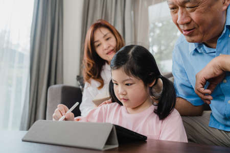 Asian grandparents and granddaughter video call at home. Senior Chinese, grandpa and grandma happy with girl using mobile phone video call talking with dad and mom lying in living room at home.の写真素材