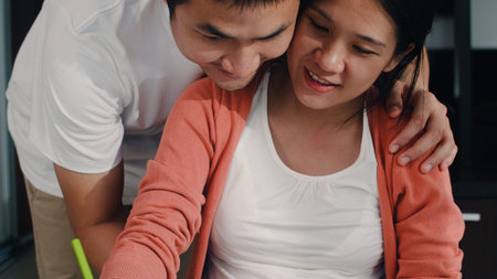 Young Asian Pregnant woman drawing baby in belly and family in notebook. Dad touch his wife belly while happy smiling positive and peaceful while take care child on table in living room at home.の写真素材