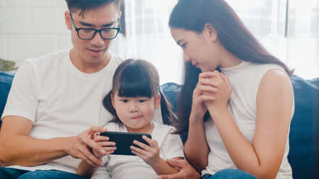 Happy Asian family dad, mom and daughter playing funny game online on smartphone sitting sofa in room at house. Self-isolation, stay at home, social distancing, quarantine for   virus prevention.の写真素材