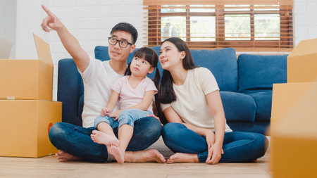 Happy Asian young family homeowners bought new house. Korean Mom, Dad, and daughter embracing looking forward to future in new home after moving in relocation sitting on floor with boxes together.の写真素材