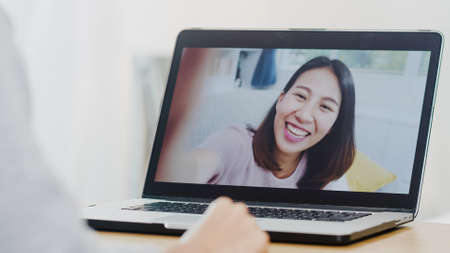 Young Asian business female using laptop video call talking with friends while working from home at bed room. Self-isolation, social distancing, quarantine for virus in new normal concept.の写真素材