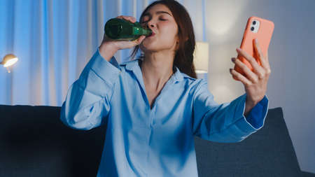 Young Asian lady drinking beer having fun happy moment night party event online celebration via video call in living room at home at night. Social distancing, quarantine for coronavirus prevention.の写真素材