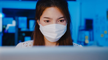 Happy Asia businesswoman wearing medical face mask for social distancing in new normal situation for virus prevention while using laptop back at work in office night. Life and work after coronavirus.の写真素材