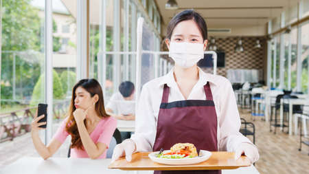 Young Asia female restaurant staff wearing protective face mask holding food tray to serving meal to customer with customer in background.の写真素材