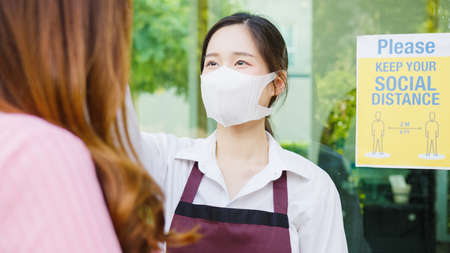 Young Asia female restaurant staff wearing protective face mask using infrared thermometer checker or temperature gun on customer's forehead before enter.の写真素材