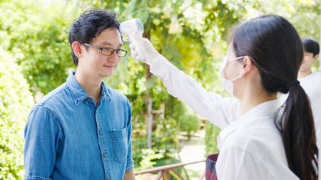 Young Asia female restaurant staff wearing protective face mask using infrared thermometer checker or temperature gun on customer's forehead before enter.の写真素材