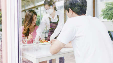 Young Asia female restaurant staff wearing protective face mask and gloves serving food to customer in cafe. Lifestyle new normal restaurant after virus and social distancing concept.の写真素材