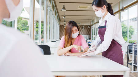 Young Asia female restaurant staff wearing protective face mask and gloves serving food to customer in cafe. Lifestyle new normal restaurant after   virus and social distancing concept.の写真素材