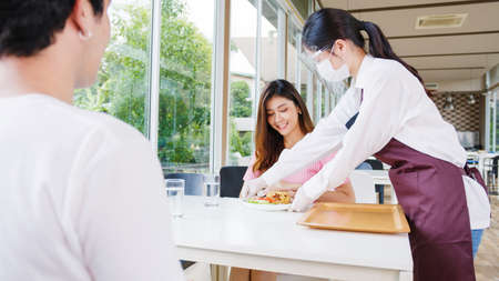 Young Asia female restaurant staff wearing protective face mask and gloves serving food to customer in cafe.の写真素材
