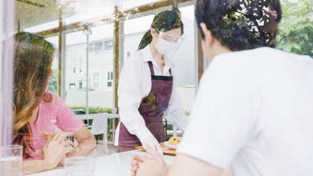 Young Asia female restaurant staff wearing protective face mask and gloves serving food to customer in cafe. Lifestyle new normal restaurant after  virus and social distancing concept.の写真素材