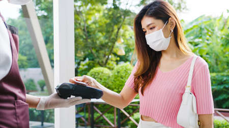 Young Asia female customer wear face mask making contactless credit card payment and take out food bag order from restaurant staff in coffee shop. Lifestyle new normal after corona virus concept.の写真素材