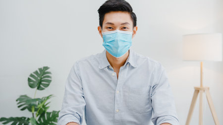 Asia businessman wear face mask social distancing in situation for virus prevention looking at camera smiling under surgical mask back to work in office workspace.の写真素材