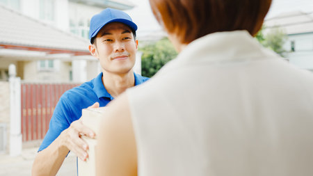 Young Asia postal delivery courier man in blue shirt handling parcel boxes for sending to customer at house and Asian female receive delivered package outdoors. Package shopping food delivery concept.の写真素材