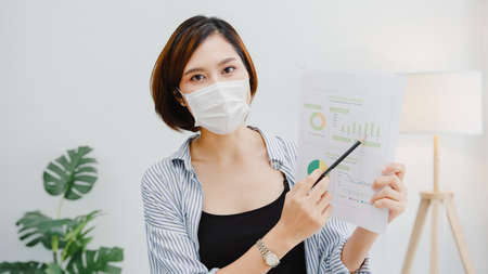 Asia businesswoman wear face mask social distancing in situation for virus prevention looking at camera presentation to colleague about plan in video call work in office.の写真素材