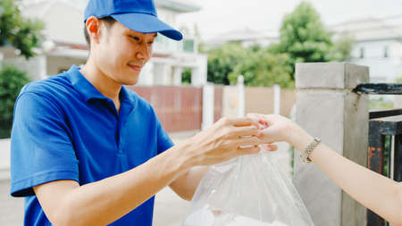 Young Asia postal delivery courier man in blue shirt handling food boxes for sending to customer at house and Asian female receive delivered package outdoors. Package shopping food delivery concept.の写真素材