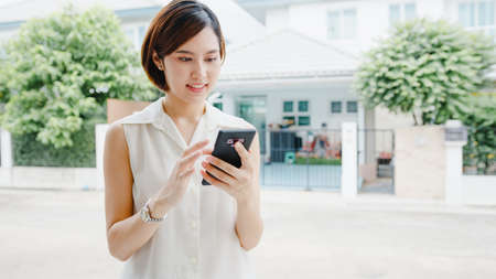 Attractive young Asia businesswoman using mobile phone checking social media internet, chatting with friends outside on street in city.の写真素材