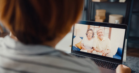 Young Asia girl using laptop video call meeting talk with elderly family dad and mom while work from home sitting on sofa at living room. Social distancing, quarantine for corona virus prevention.の写真素材