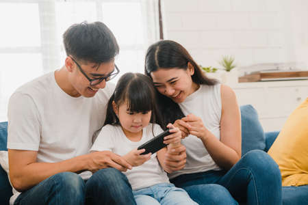 Happy Asian family dad, mom and daughter playing funny game online on smartphone sitting sofa in room at house. Self-isolation, stay at home, social distancing, quarantine for virus prevention.の写真素材