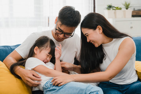 Happy cheerful Asian family dad, mom and daughter having fun cuddling playing on sofa while birthday at house. Self-isolation, stay at home, social distancing, quarantine for  virus prevention.の写真素材