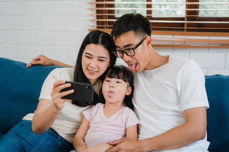 Happy cheerful Asian family dad, mom and kids having fun and using smart phone video call on sofa at house. Self-isolation, stay at home, social distancing, quarantine for virus prevention.の写真素材
