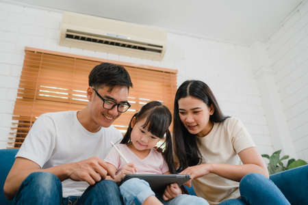 Happy Asian family dad, mom and daughter using computer tablet technology sitting sofa in living room at house. Self-isolation, stay at home, social distancing, quarantine for virus prevention.の写真素材