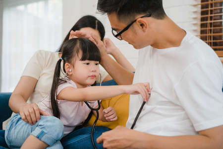 Happy cheerful Asian family dad, mom and daughter playing funny game as doctor having fun on sofa at home.の写真素材