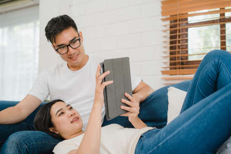 Asian couple using tablet for online shopping in internet in living room at home, sweet couple enjoy love moment while lying on the sofa when relax at home. Lifestyle couple relax at home concept.の写真素材