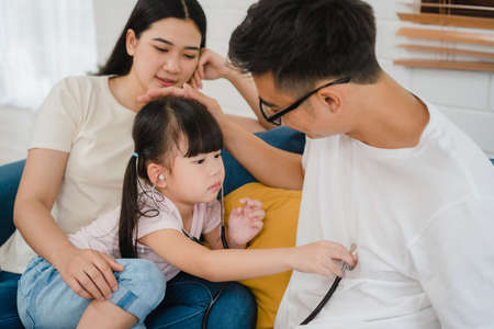 Happy cheerful Asian family dad, mom and daughter playing funny game as doctor having fun on sofa at home.の写真素材