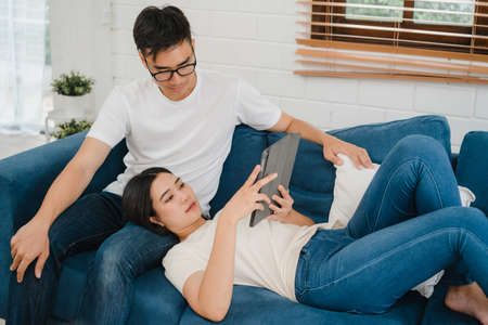 Asian couple using tablet for online shopping in internet in living room at home, sweet couple enjoy love moment while lying on the sofa when relax at home. Lifestyle couple relax at home concept.の写真素材