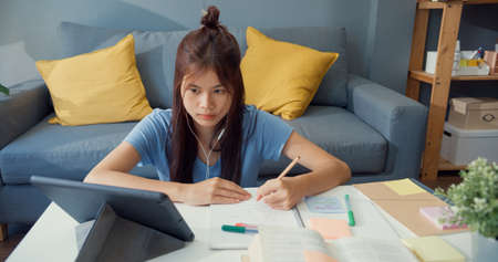 Young Asia girl teenager with casual wear headphones use digital tablet learn online write lecture notebook in living room at house. Isolate education online e-learning coronavirus pandemic concept.の写真素材
