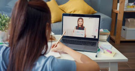 Young Asia girl with casual use computer laptop video call learn online with teacher write lecture notebook living room at house. Isolate education online e-learning coronavirus pandemic concept.の写真素材